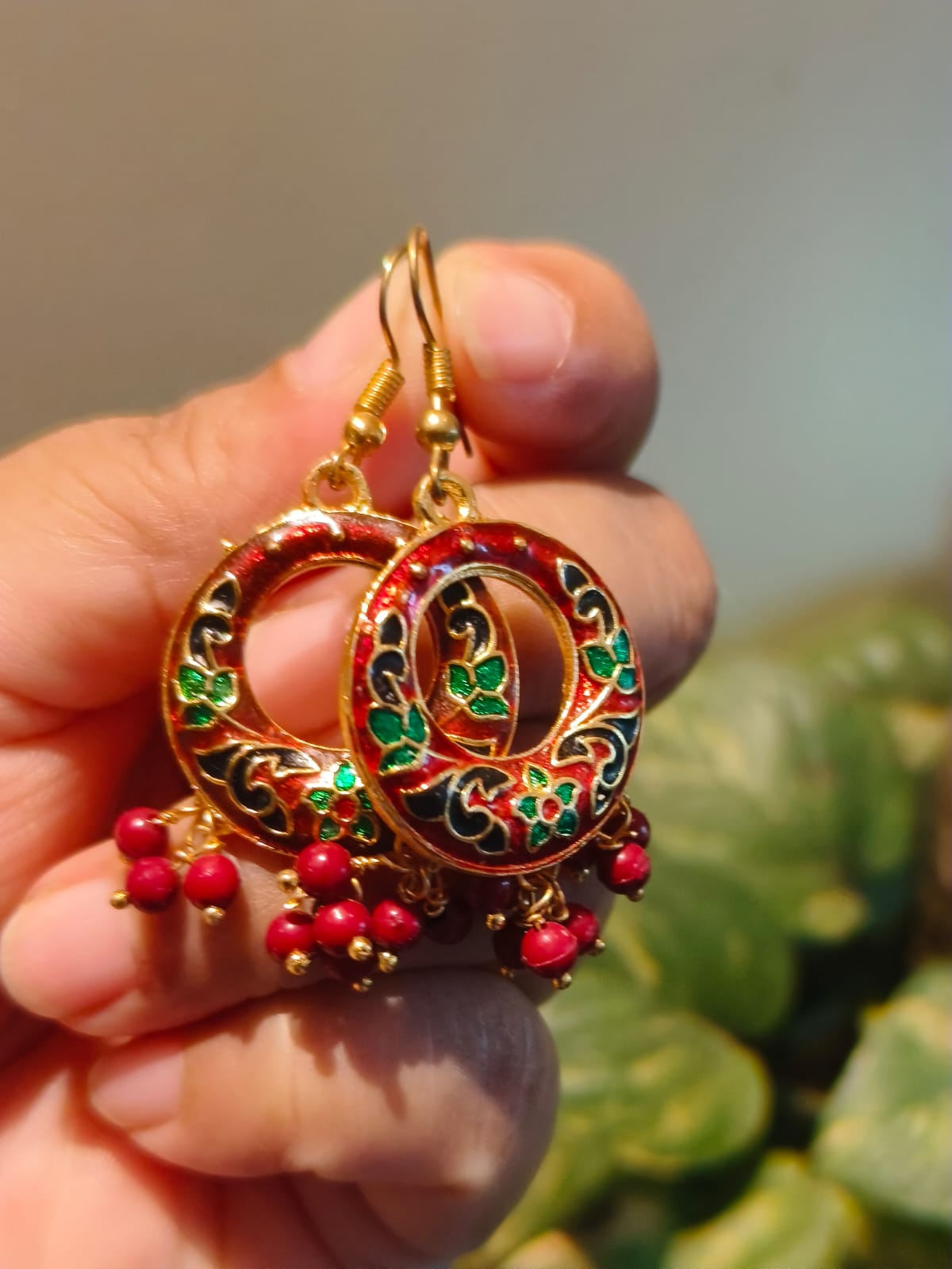 Close-up of handcrafted red Meenakari chandbali earrings with green and black enamel floral motifs, gold-toned base and ruby-red bead drops, held in hand | Rivayat The Heritage