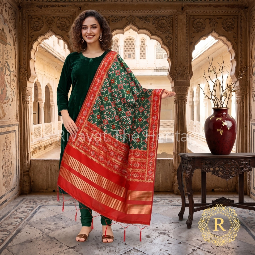 A female model with curly hair showcasing the red and green Rivayat Navdurga Patola Silk Dupatta, elegantly draped over a rich green velvet kurta to create a luxury contrast. The background features a realistic, traditional Indian palace setting with intricately carved stone archways, marble flooring, and an ornate wooden table displaying a decorative maroon vase.